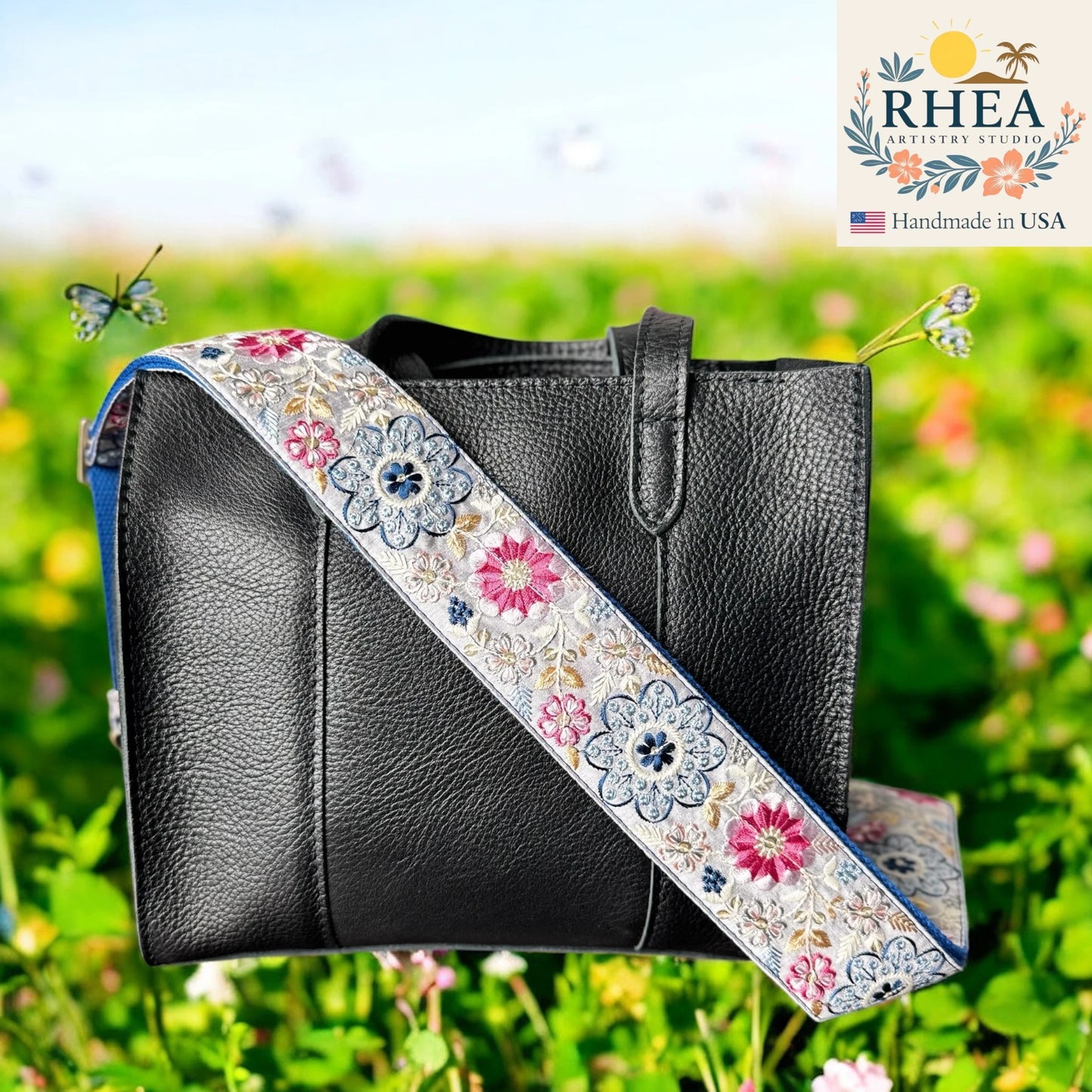 Black handbag with a floral purse strap against a blurred green outdoor background, featuring the RHEA Artistry Studio logo.