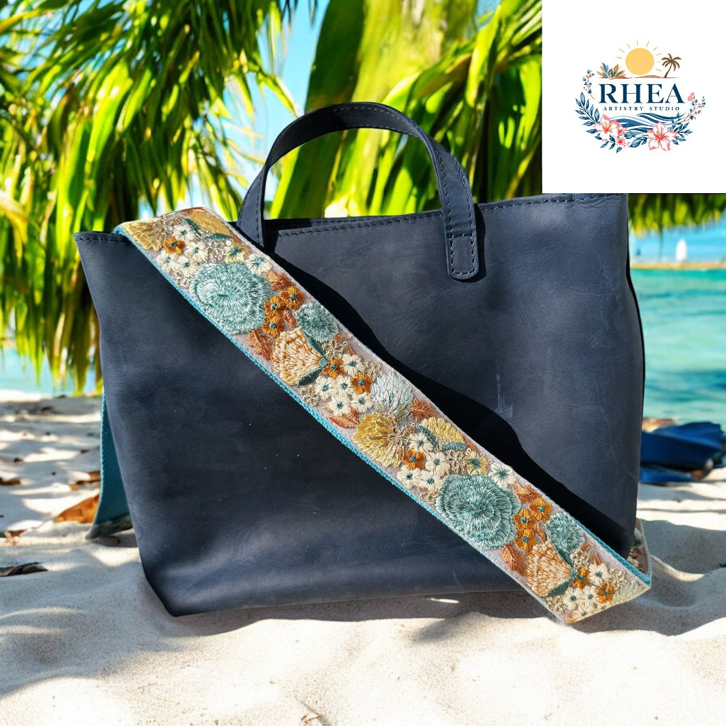 A navy leather handbag with a wide strap embroidered in muted teal, gold, and rust floral patterns, photographed in a lush garden with soft green and pink blooms.