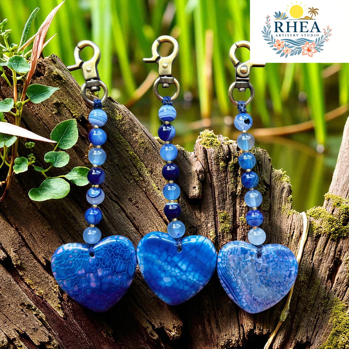 Blue heart-shaped keychains with beads on a wooden log with greenery in the background, featuring the RHEA brand.
