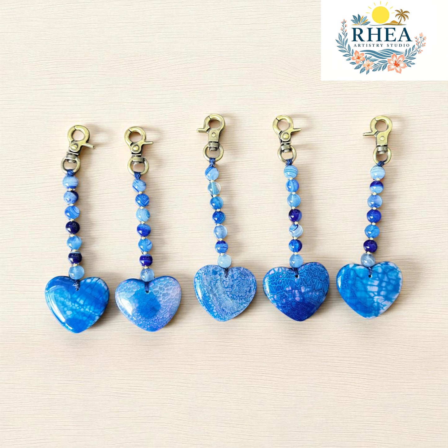 Five blue heart-shaped keychains with beaded charms on a beige background, featuring the RHEA brand logo.