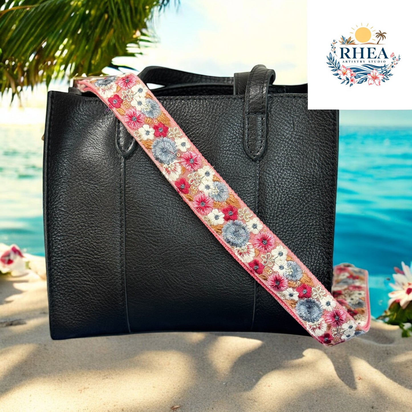 A handcrafted embroidered strap featuring intricate floral patterns in coral, blue, and white rests on a black leather bag. The photo is set against a tropical beach backdrop with the ocean, palm trees, and scattered flowers, evoking a sense of warmth, artistry, and wanderlust.