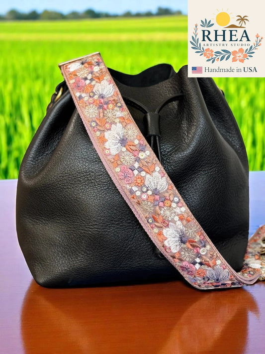 Black handbag with a floral leather bag strap against a green field background, featuring the RHEA brand.