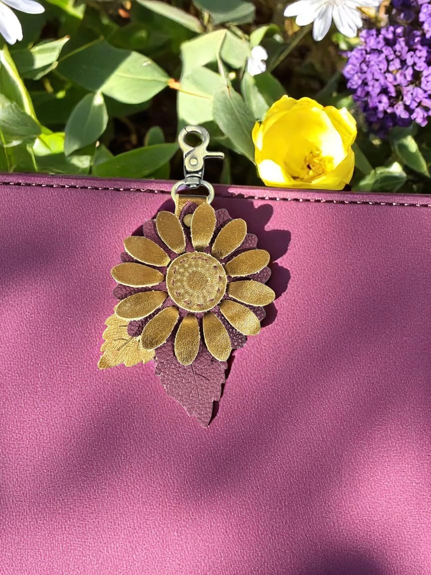 Metallic gold and plum leather flower charm with layered leaf detail in a garden setting on a plum leathered handbag