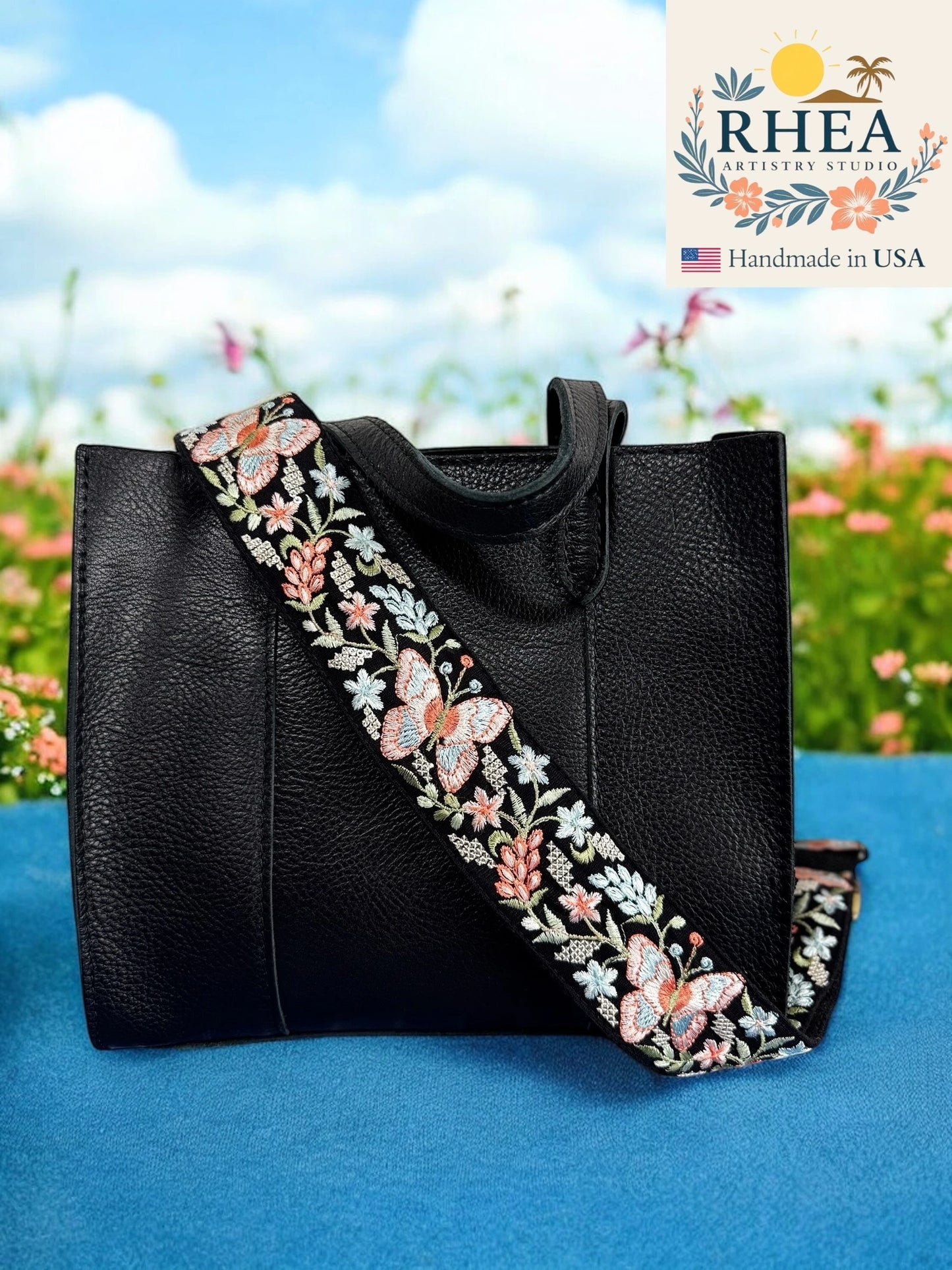 Black handbag with a floral purse strap on a blue surface with a blurred natural background, featuring the RHEA Artistry Studio logo.
