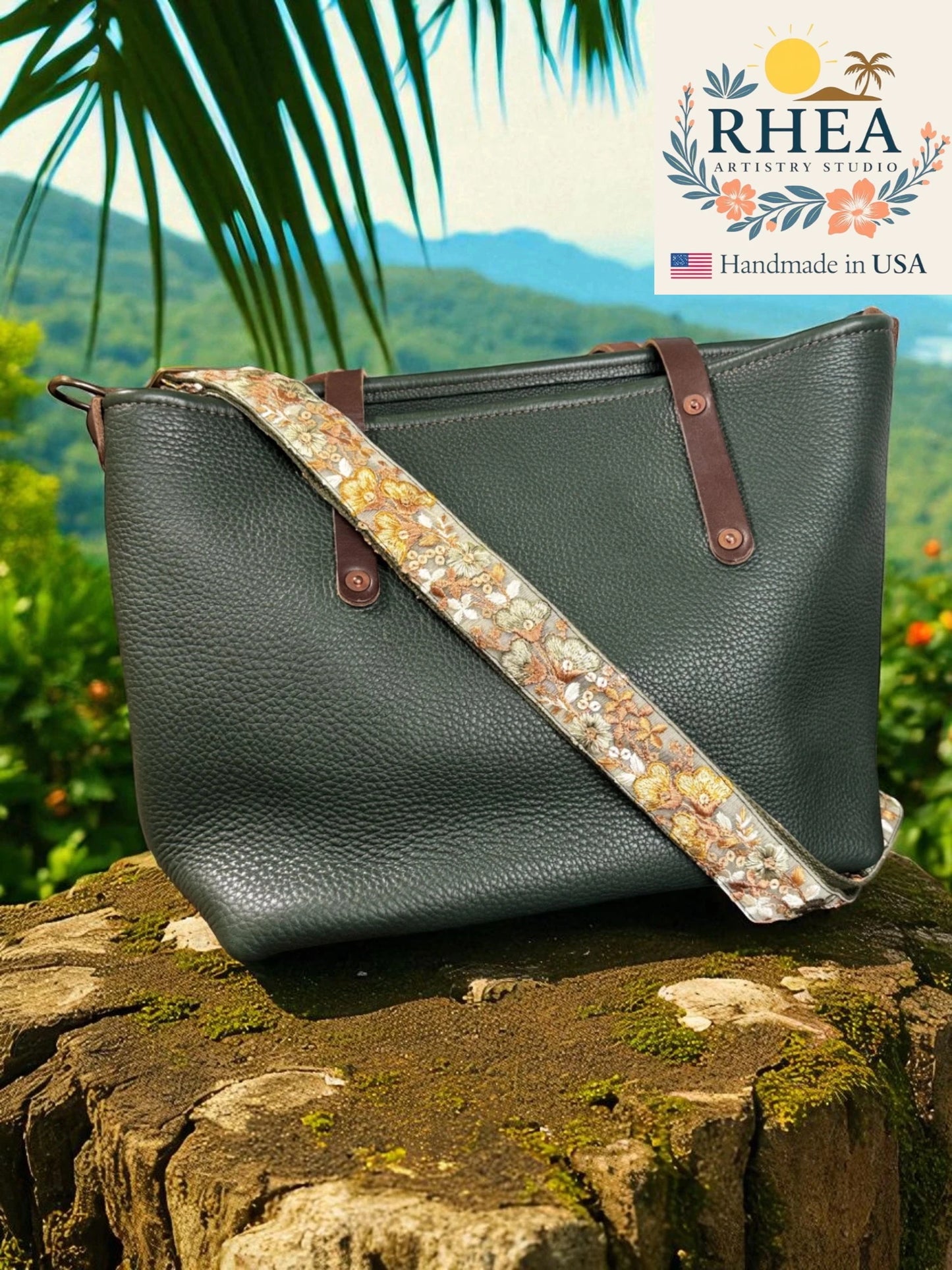 Dark green leather handbag with floral purse strap on a stone surface, with a scenic background and RHEA branding.