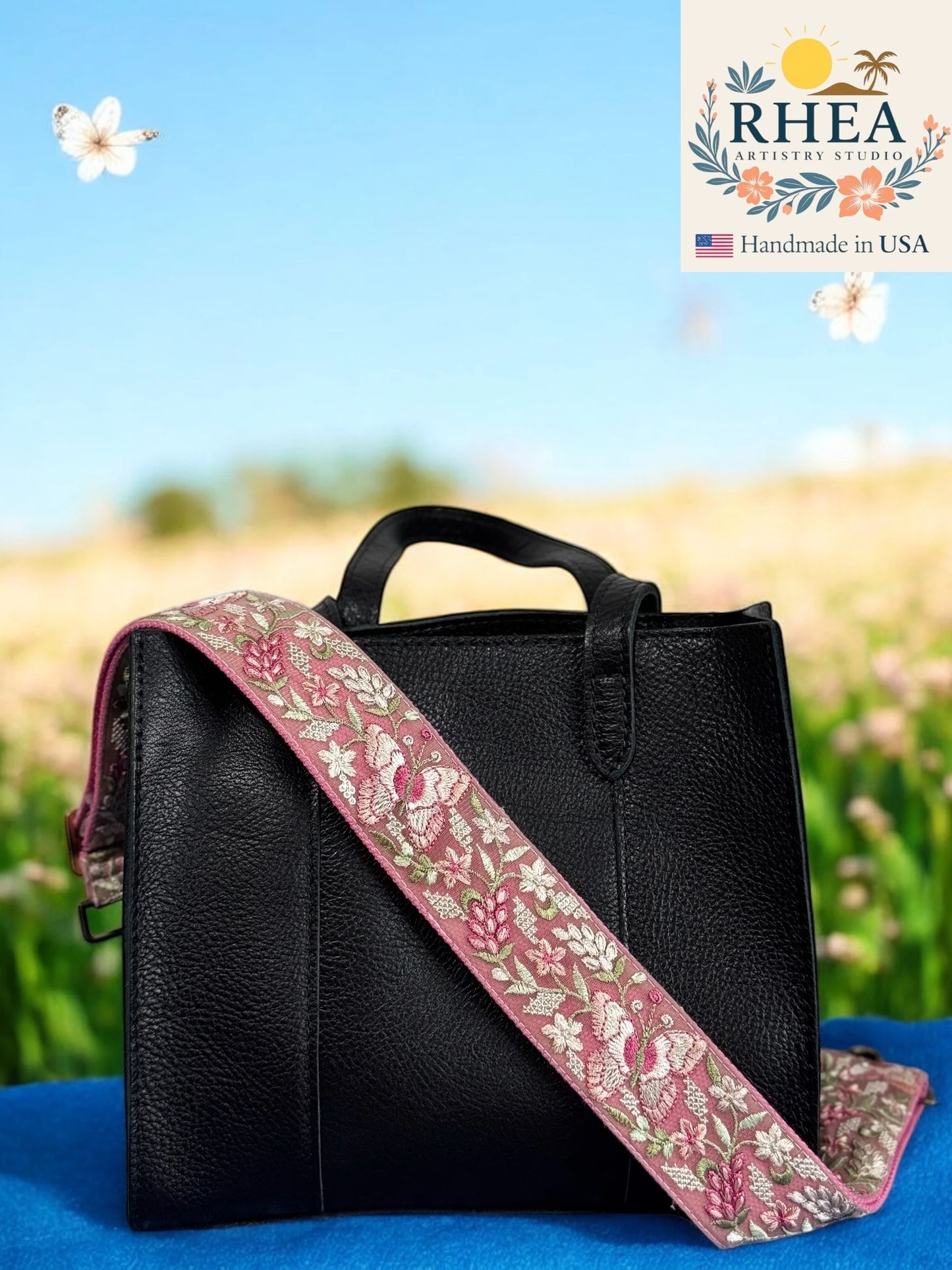 Black handbag with a floral purse strap on a blue surface with a blurred natural background, featuring the Rhea Artisan Studio logo.