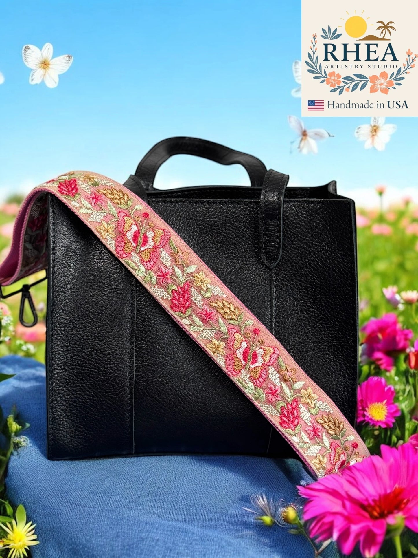 Black handbag with a floral purse strap on a blue surface with flowers in the background, featuring the RHEA brand.