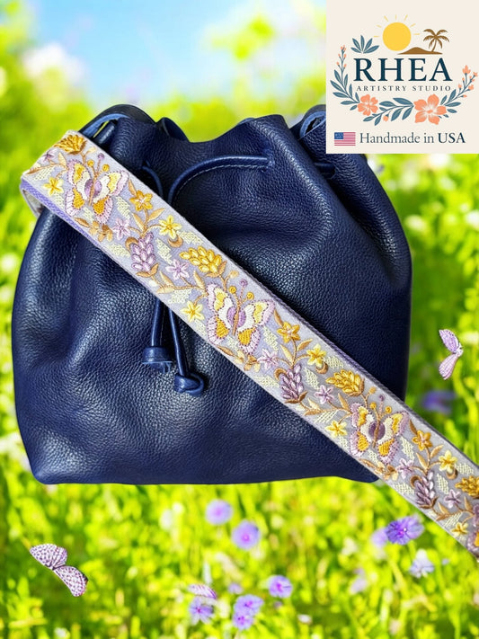 Navy blue leather bag with a decorative purse strap on a grassy background, featuring the RHEA brand.