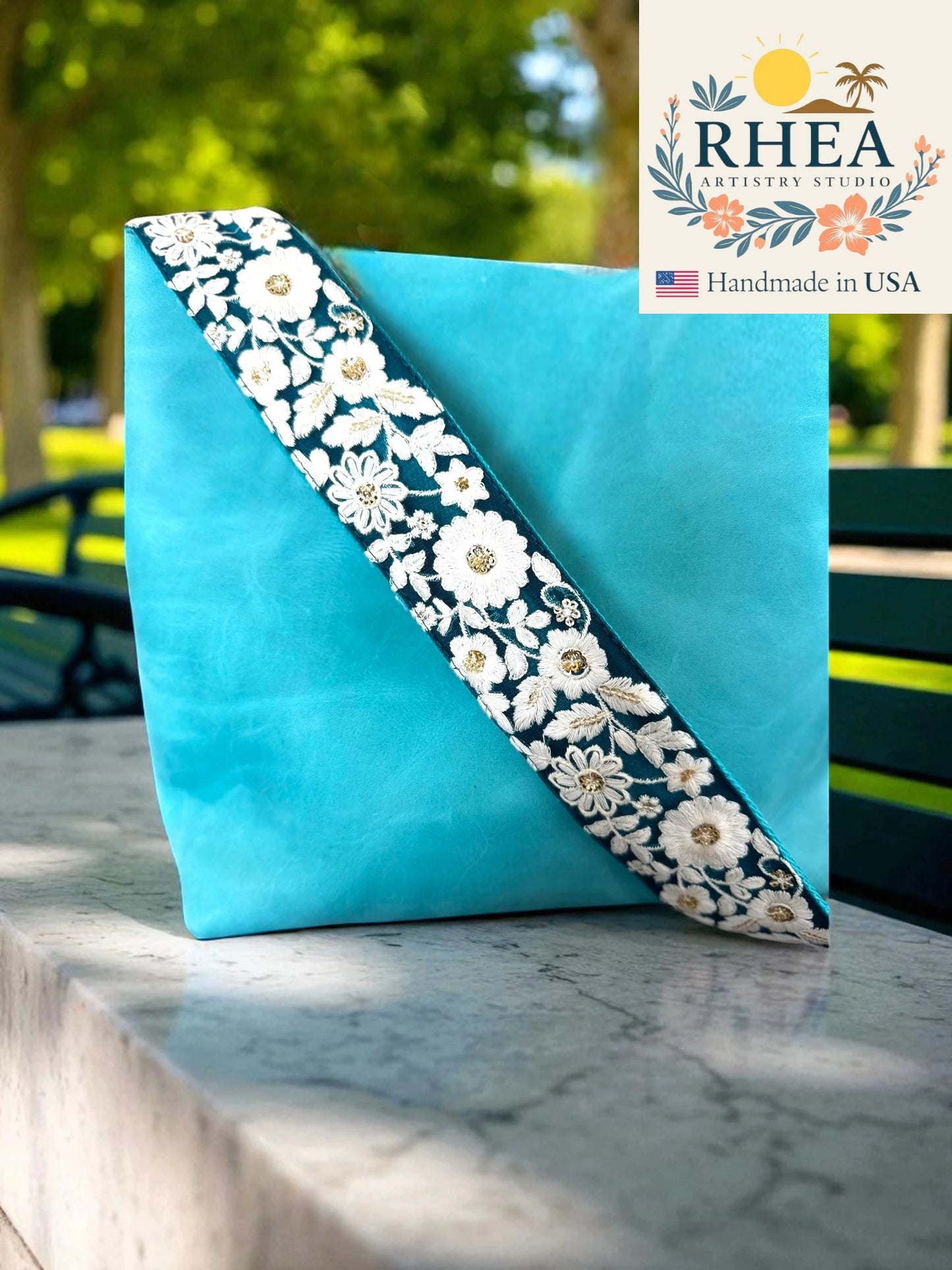 Turquoise clutch bag with floral embroidery purse strap on a marble surface, RHEA Artistry Studio logo visible.