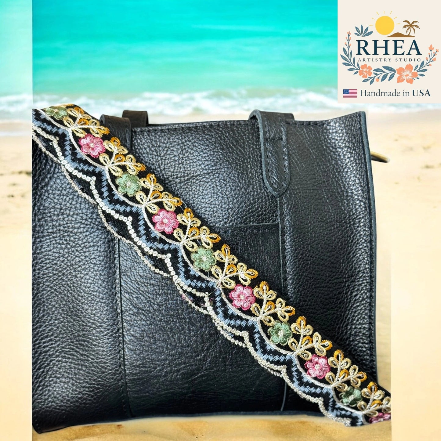 Black leather handbag with colorful floral strap on a beach background, featuring the RHEEA brand.