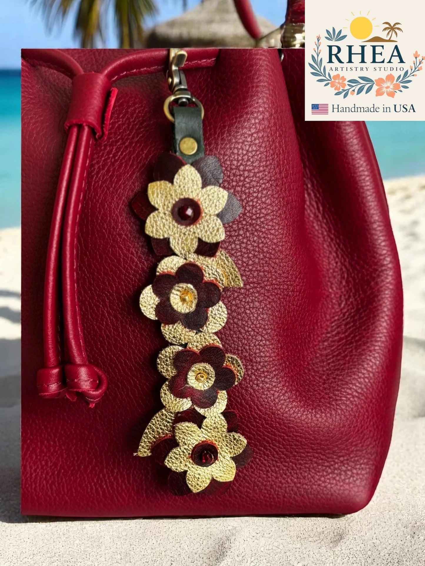 Red leather bag with floral leather bag charm on a beach, featuring RHEA brand.