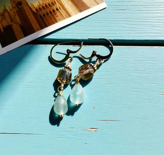 Gold earrings with green teardrop-shaped stones on a light blue wooden surface.