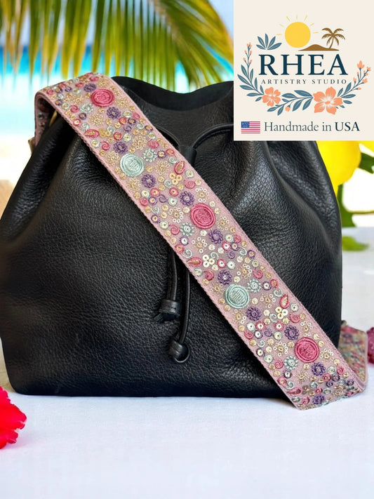 Black leather bag with a floral purse strap on a white surface, palm leaves in the background, and RHEA Artistry Studio logo.