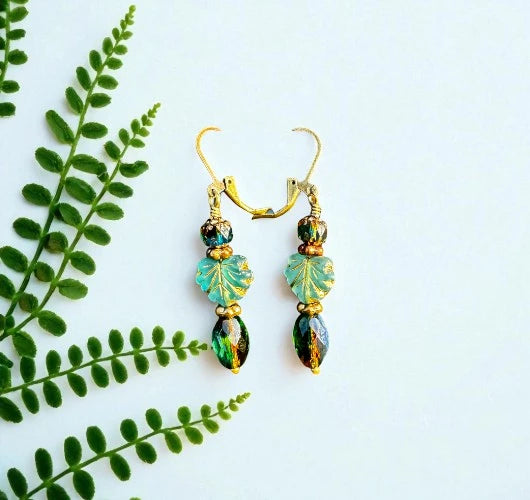 Pair of earrings with green and gold beads on a light blue background with green leaves.