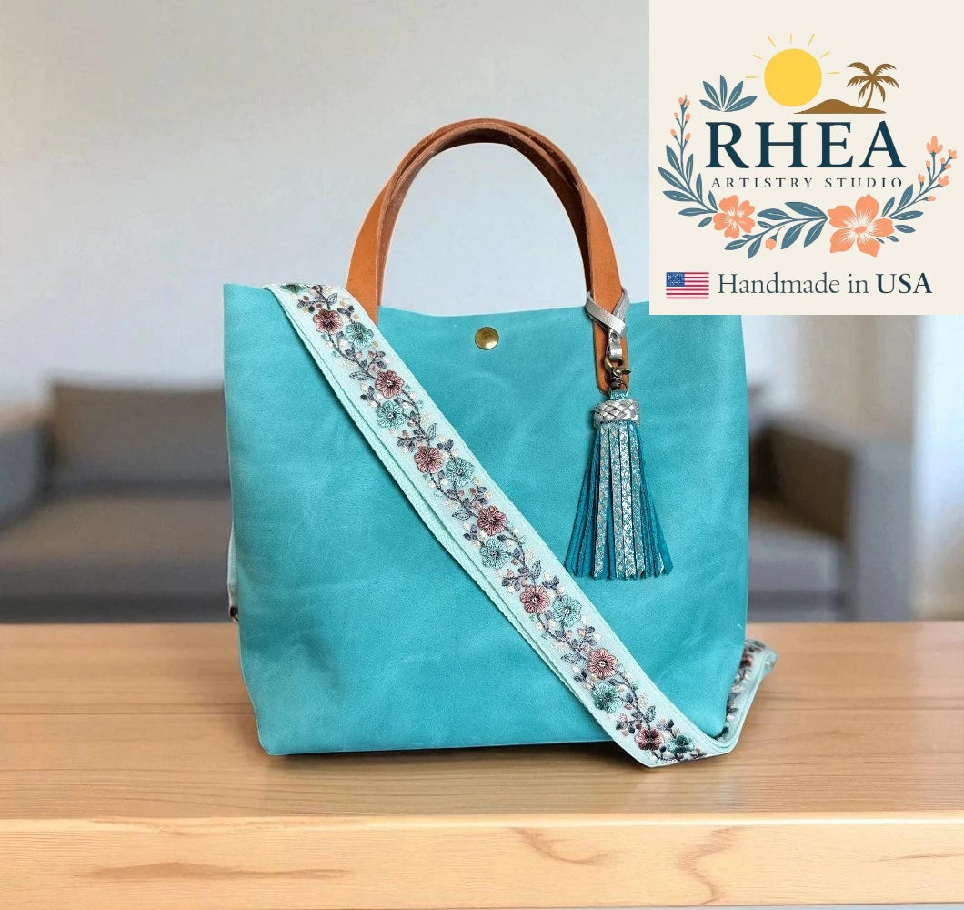 Turquoise handbag with floral bag strap and tassel on a wooden surface, RHEA Artistry Studio logo visible.