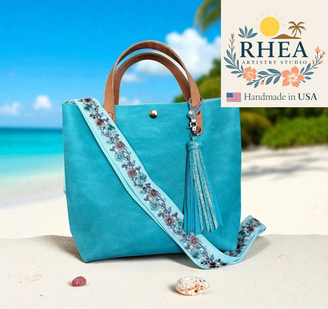 Turquoise handbag with floral bag strap and tassel on a beach, featuring RHEA Artistry Studio logo.