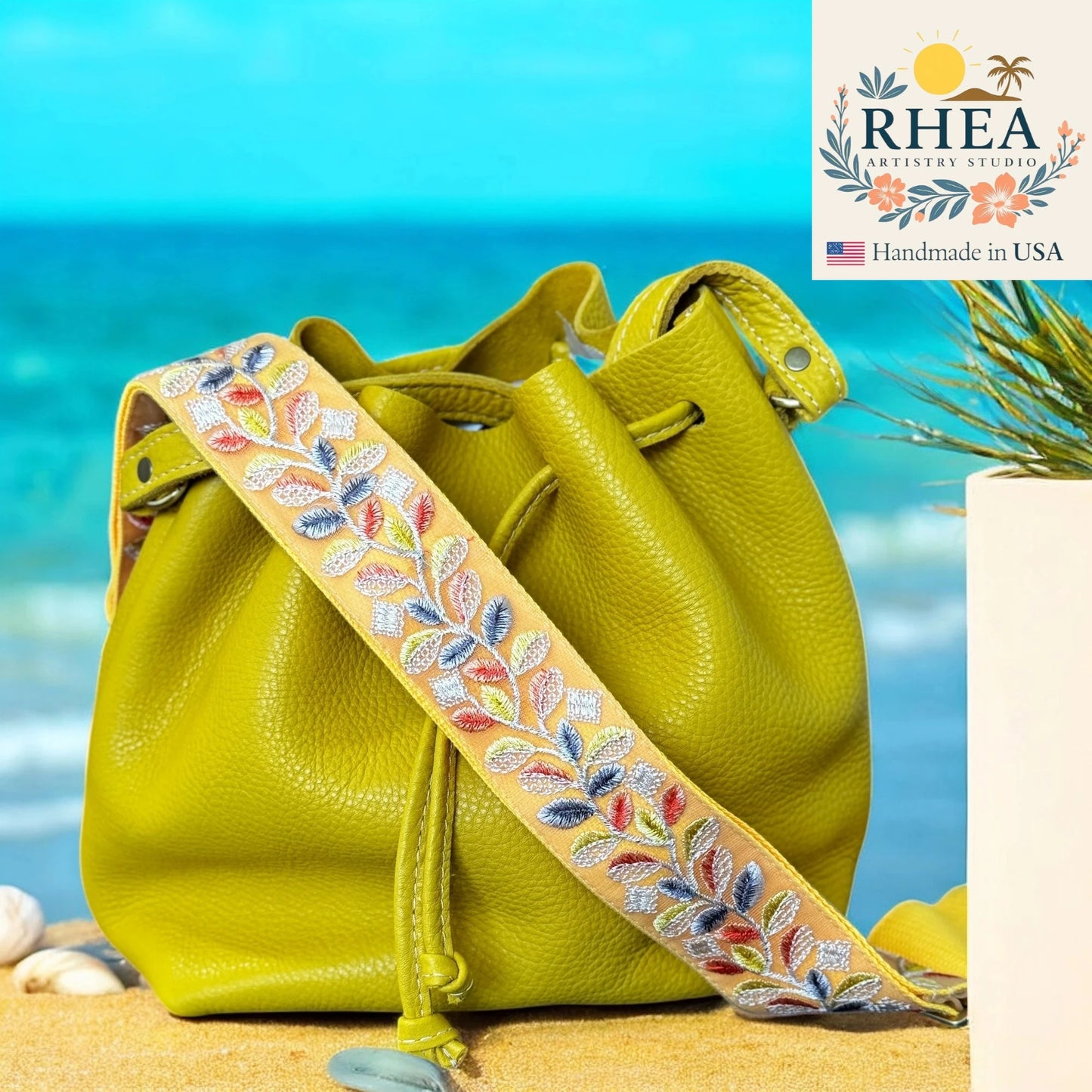 Yellow leather bag with an embroidered strap on a beach, featuring the RHEEA brand.