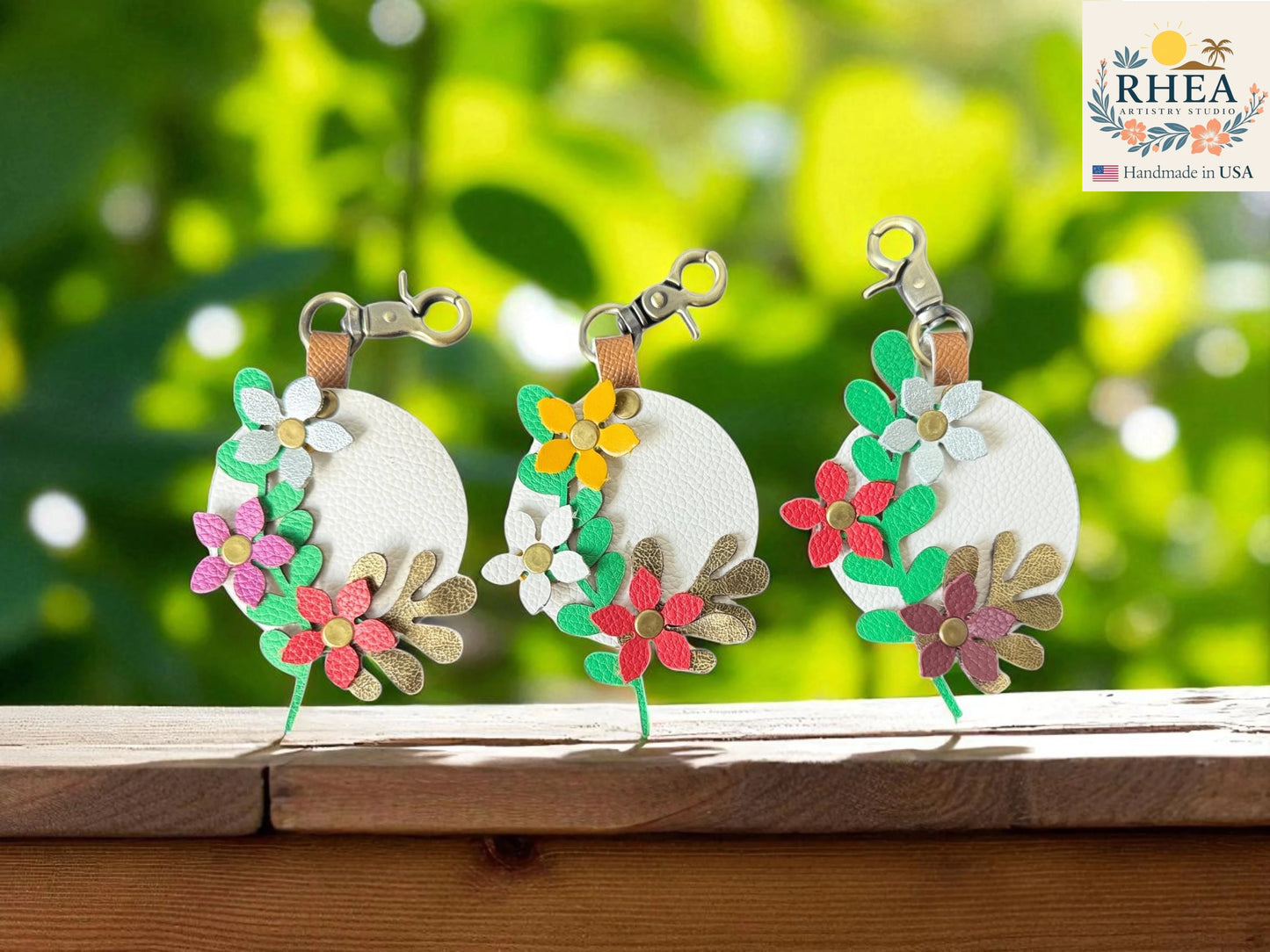 Three floral leather bag charms on a wooden surface with a blurred green background