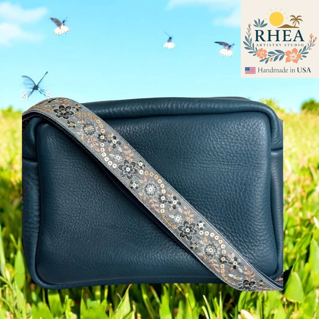 Navy blue handbag with decorative purse strap on grass with butterflies, RHEA brand logo in the corner.