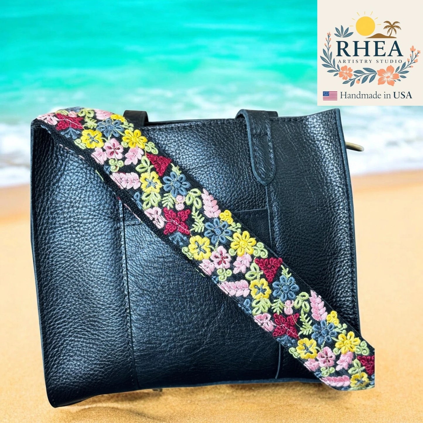 Black handbag with a colorful floral strap on a beach background, featuring the RHEA brand.