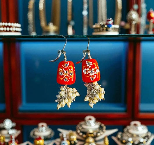 Red earrings with floral designs and pearl-like beads against a blurred jewelry store background.