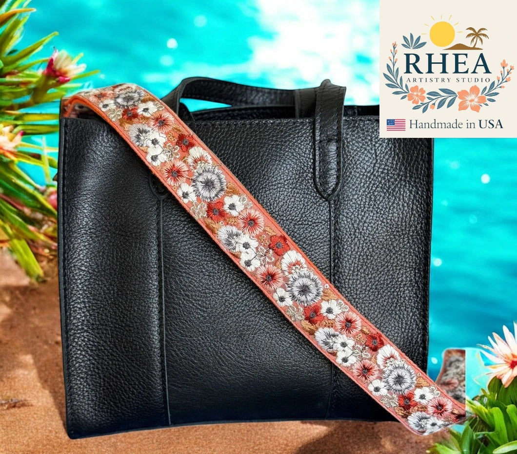 Black handbag with a floral purse strap against a bright blue background, featuring the Rhea Artistry Studio logo.