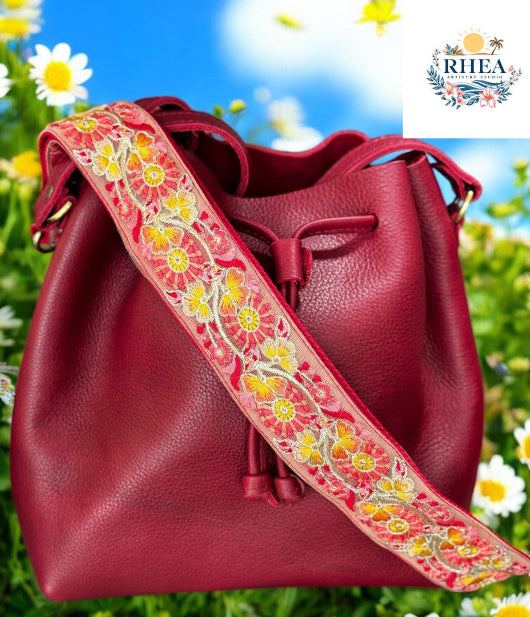 A black purse with a pink and green floral embroidered strap placed on a blue surface with a bright sky and greenery in the background.