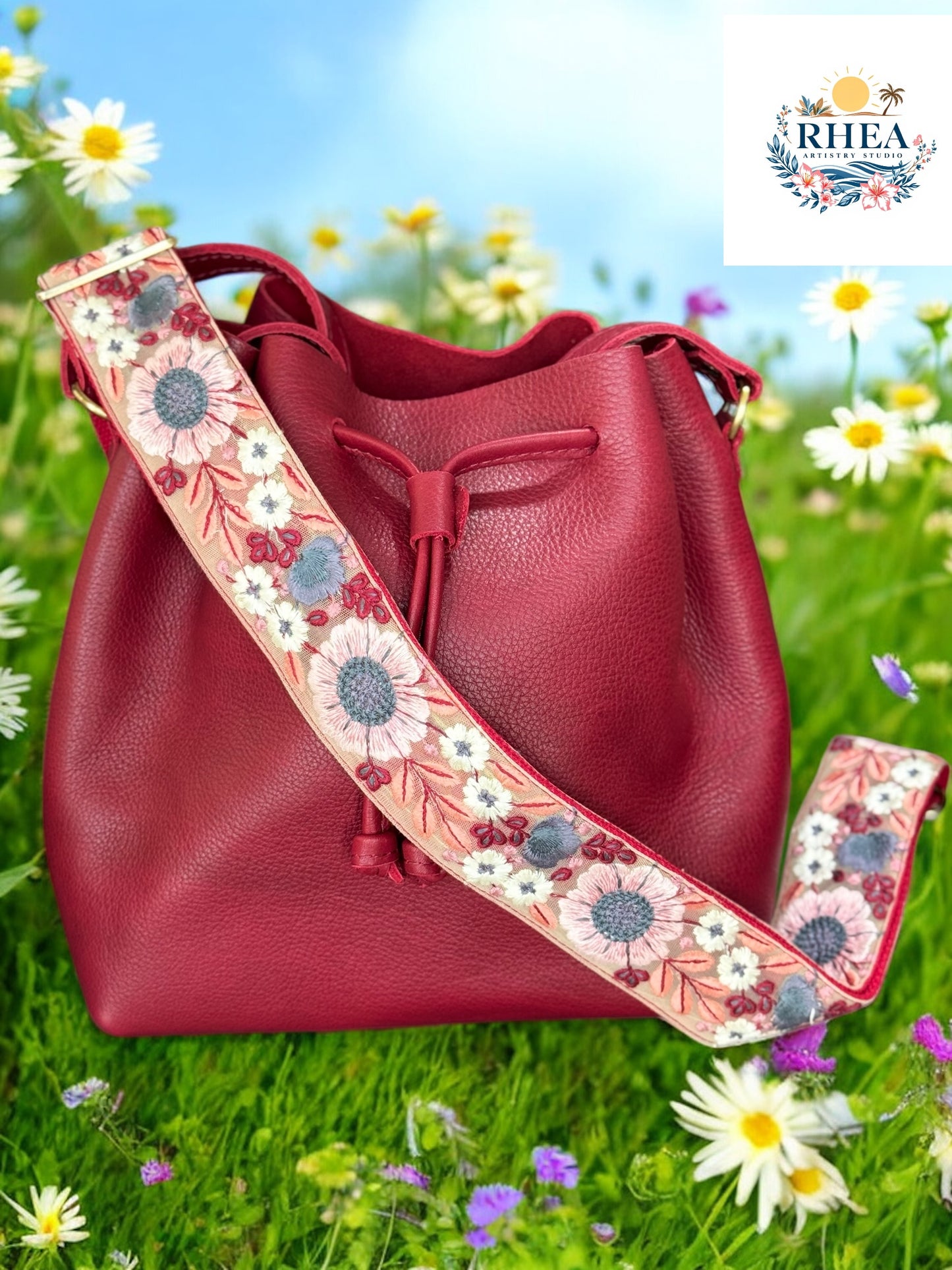 Handcrafted red crossbody bag strap with intricate floral embroidery in pink, blue, and white, shown attached to a matching red bucket bag with garden in the background. Made in USA.