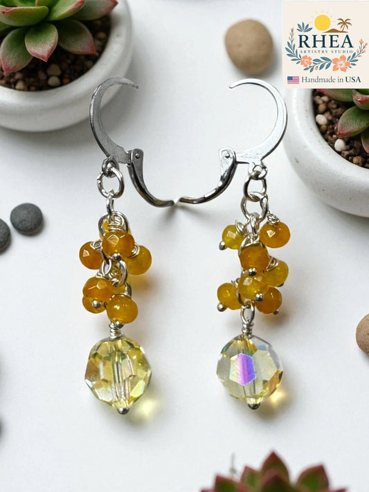 Beaded cute earrings with yellow and clear beads on a white surface with plants in the background, featuring the RHEA brand.