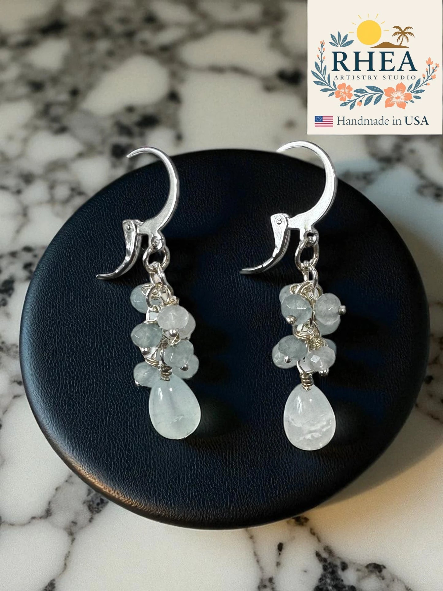 pale blue earrings for women with teardrop-shaped gemstones on a dark surface, branded 'RHEA'.