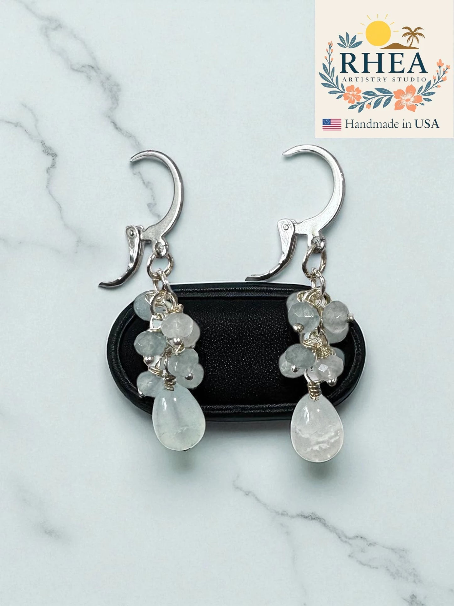 Pale blue earrings for women with clear stones on a marble surface, branded 'RHEA Artistry Studio'.