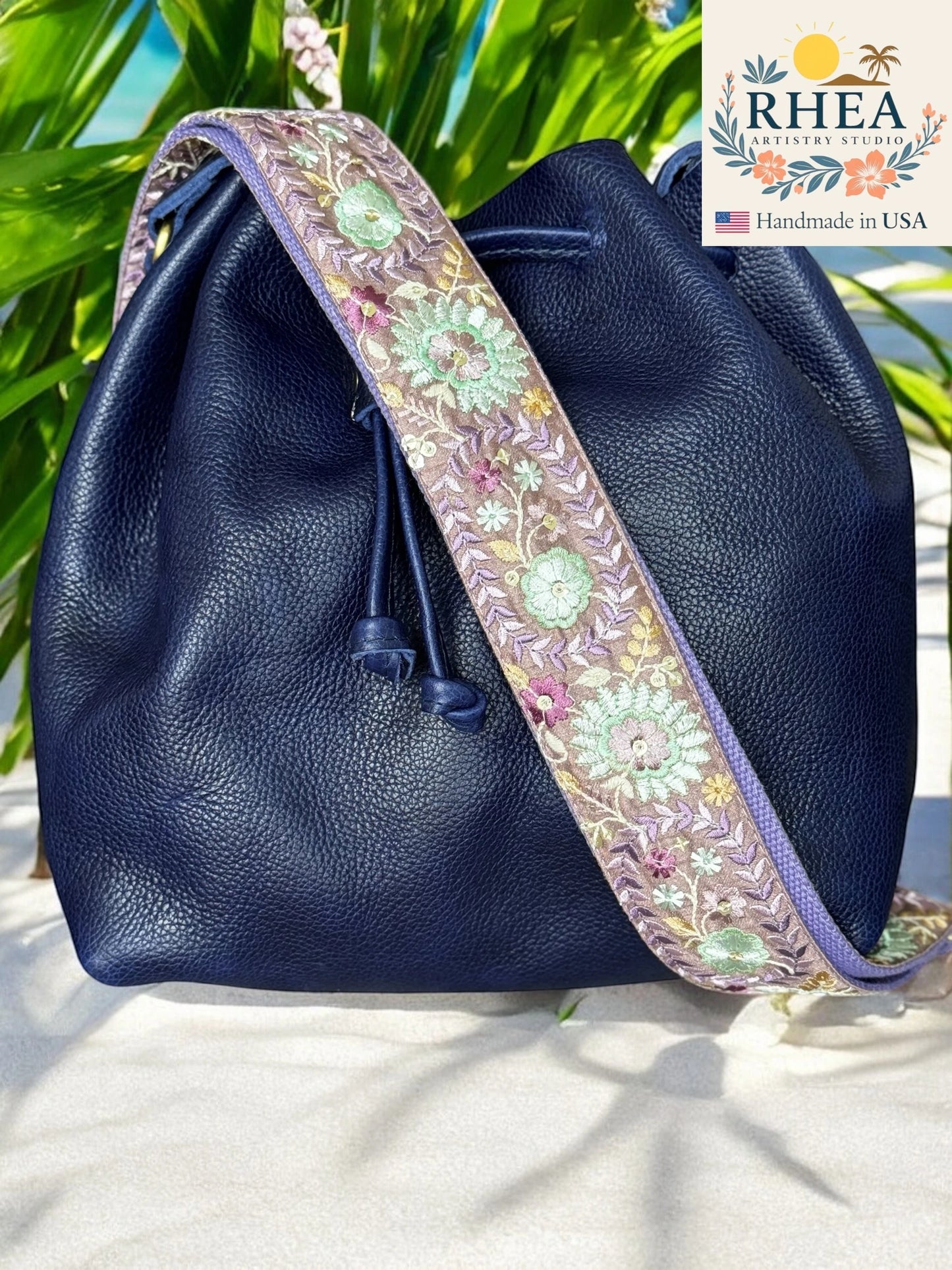 Navy blue leather bag with a floral embroidered purse strap on a white surface with greenery in the background, featuring the RHEA brand.