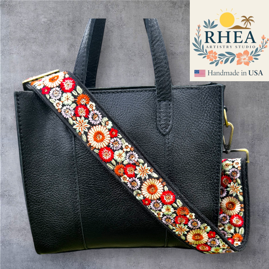 Black handbag with a colorful floral purse strap on a gray background, featuring the Rhea Artistry Studio logo.