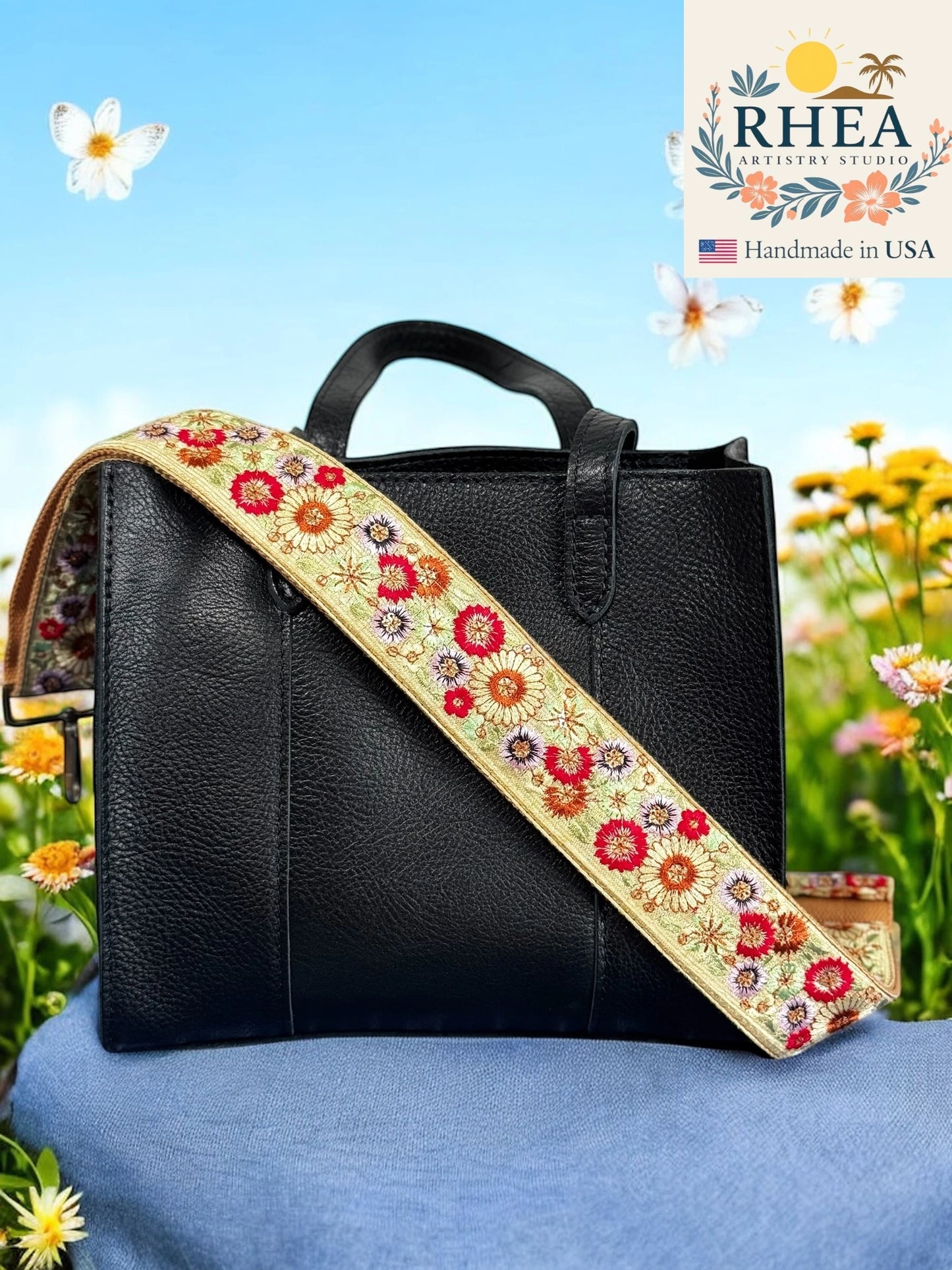 Black handbag with a floral purse strap on a blue surface with flowers in the background, featuring the Rhea brand.