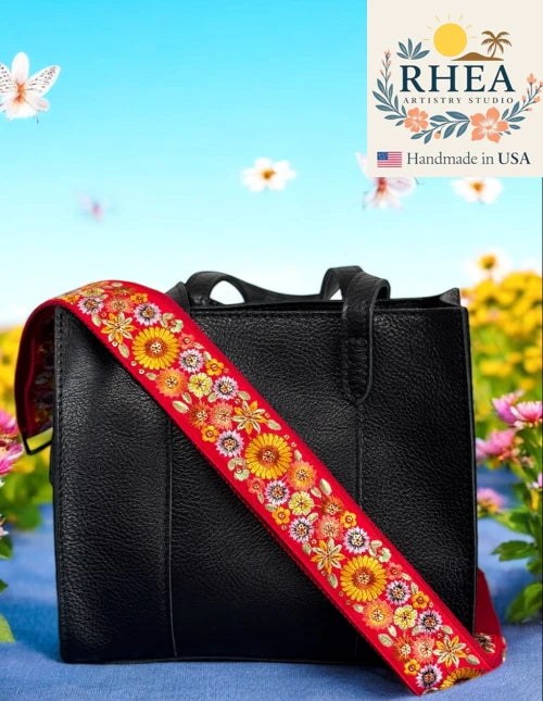 Black leather handbag with a colorful floral purse strap against a floral background, featuring the Rhea Artistry Studio logo.