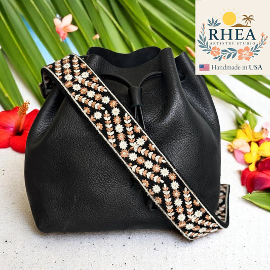 Black handbag with a decorative purse strap on a white surface with flowers and RHEA Artistry Studio logo.