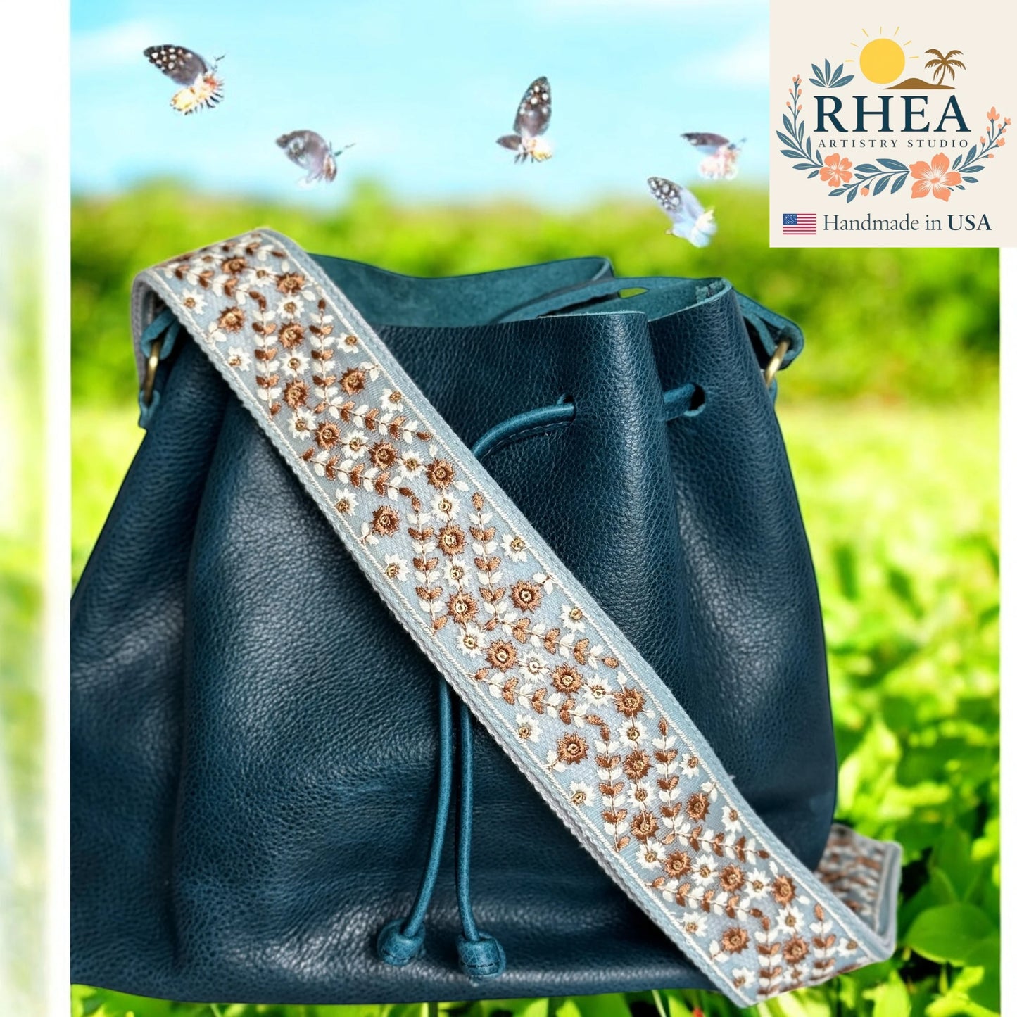 Blue leather bag with an embellished spurse strap against a green outdoor background, featuring the RHEA brand.