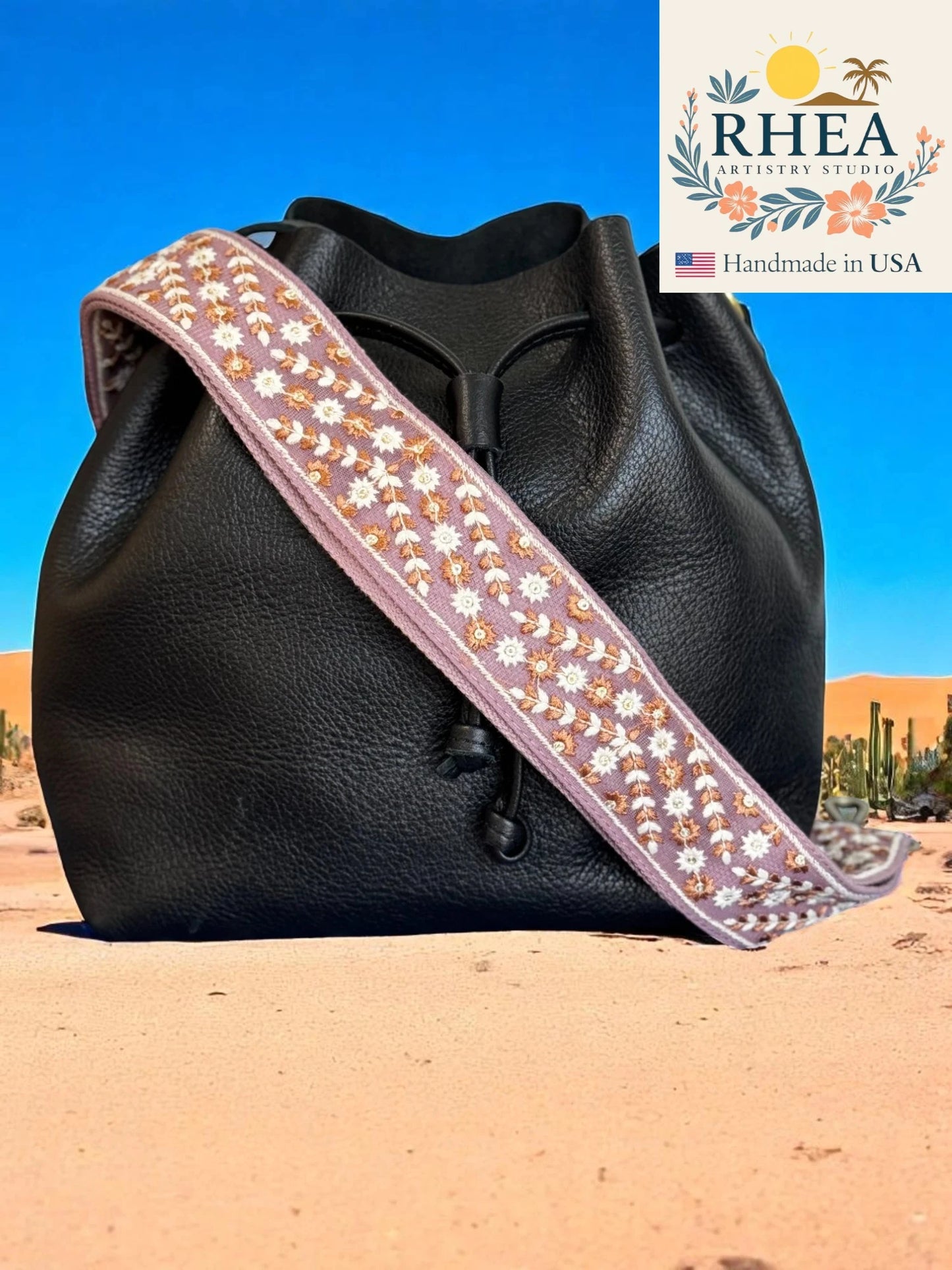 Black leather bag with a decorative purse strap against a desert background, featuring the RHEA brand.