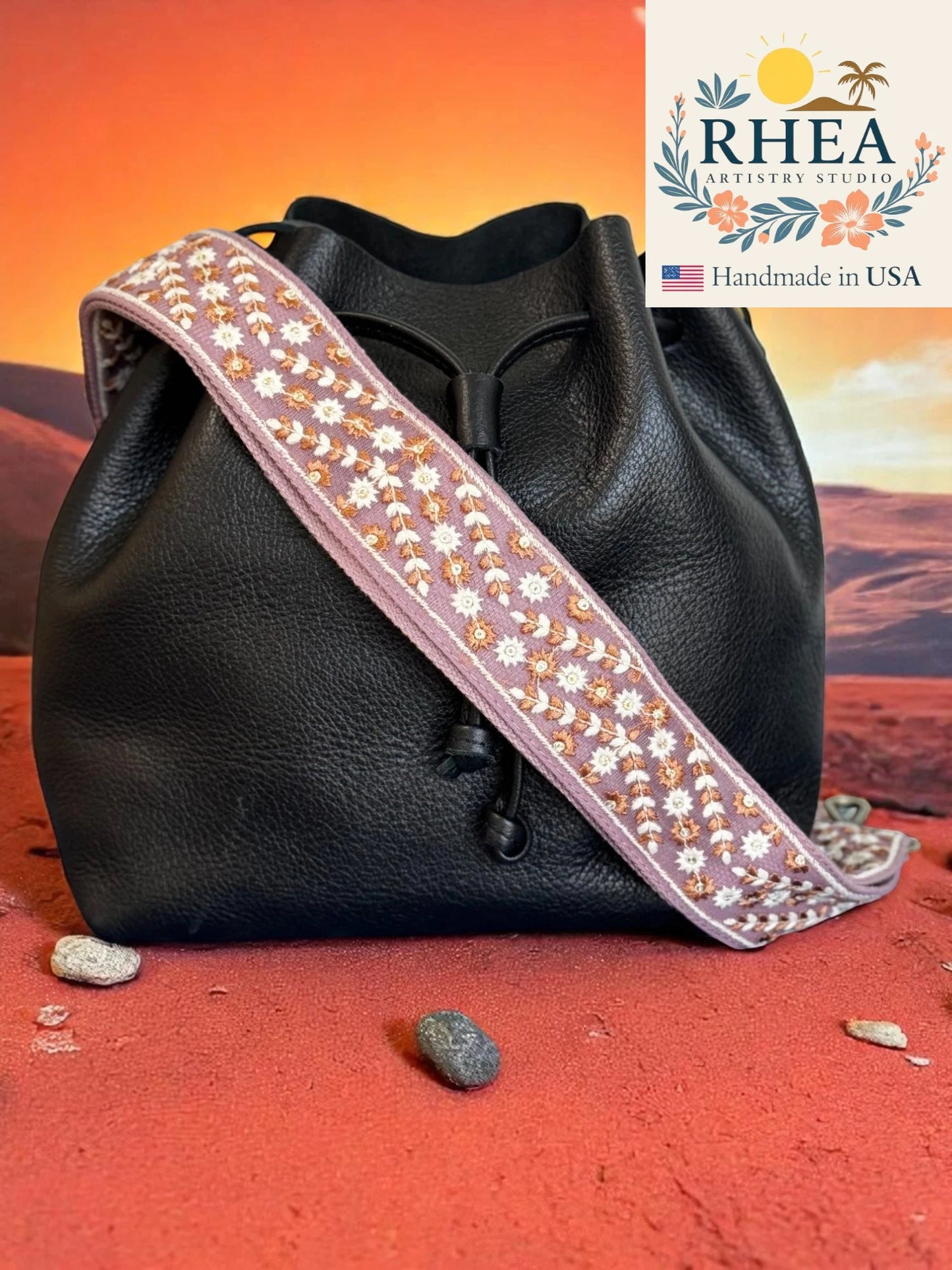 Black leather bag with a decorative purse strap on a desert landscape background, featuring the RHEA brand.