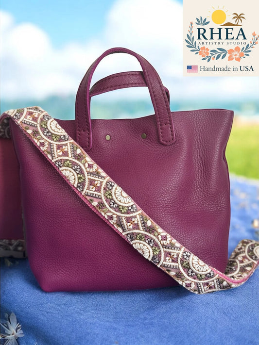 Purple handbag with a patterned purse strap on a blue surface, RHEA brand visible.