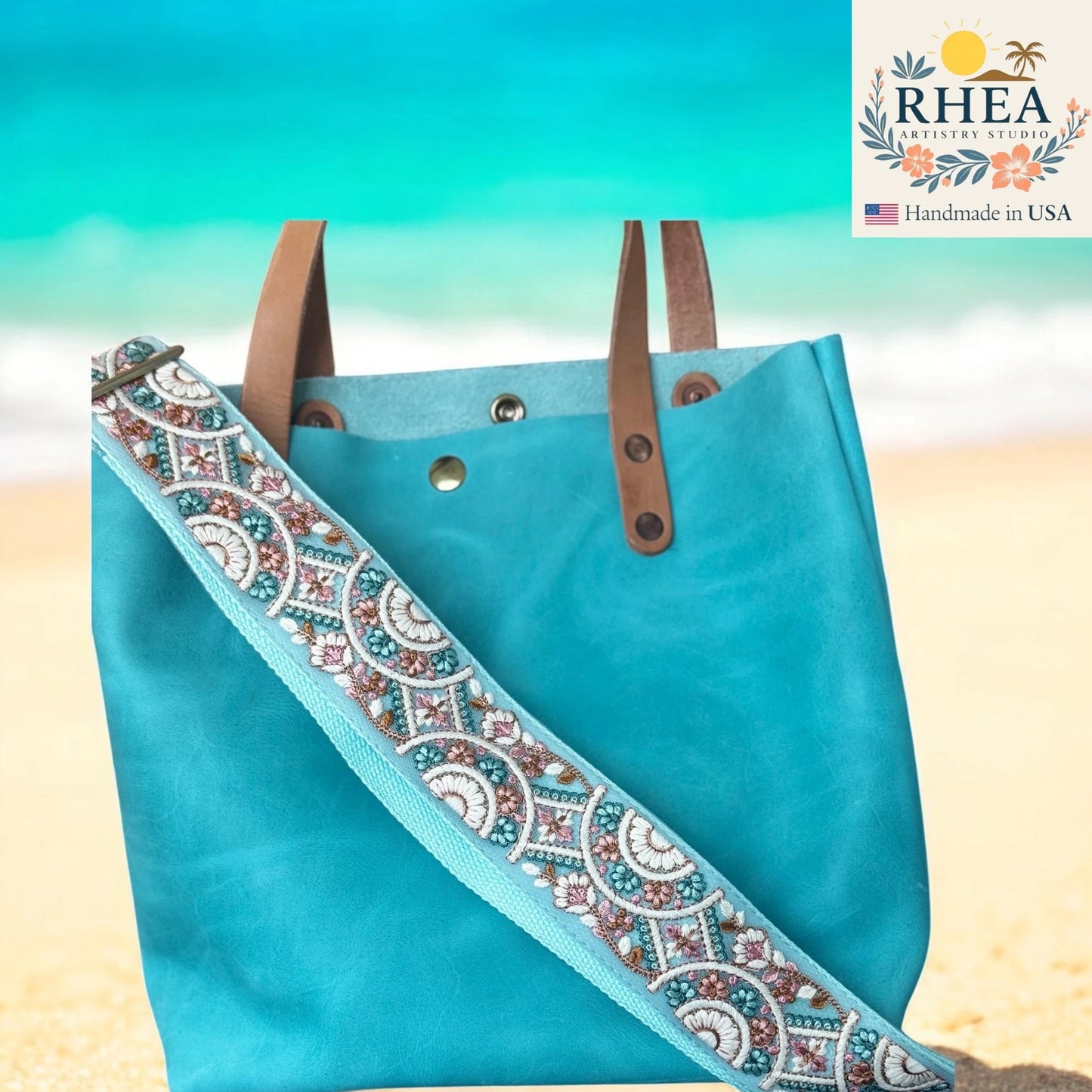 Blue tote bag with decorative purse strap on a beach background, featuring the RHEA brand.