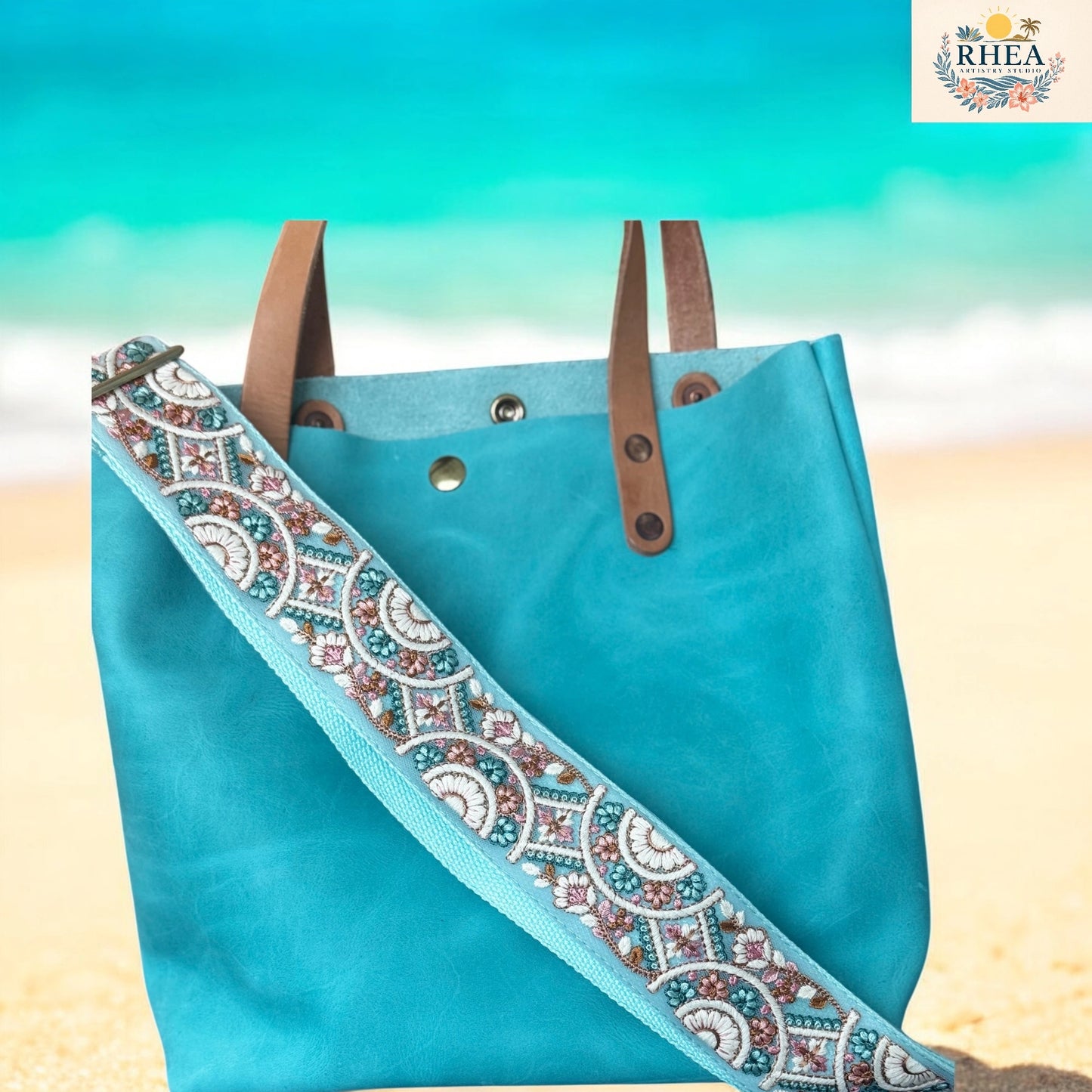 Blue tote bag with brown straps and an embroidered strap on a beach background, featuring the RHEA brand.