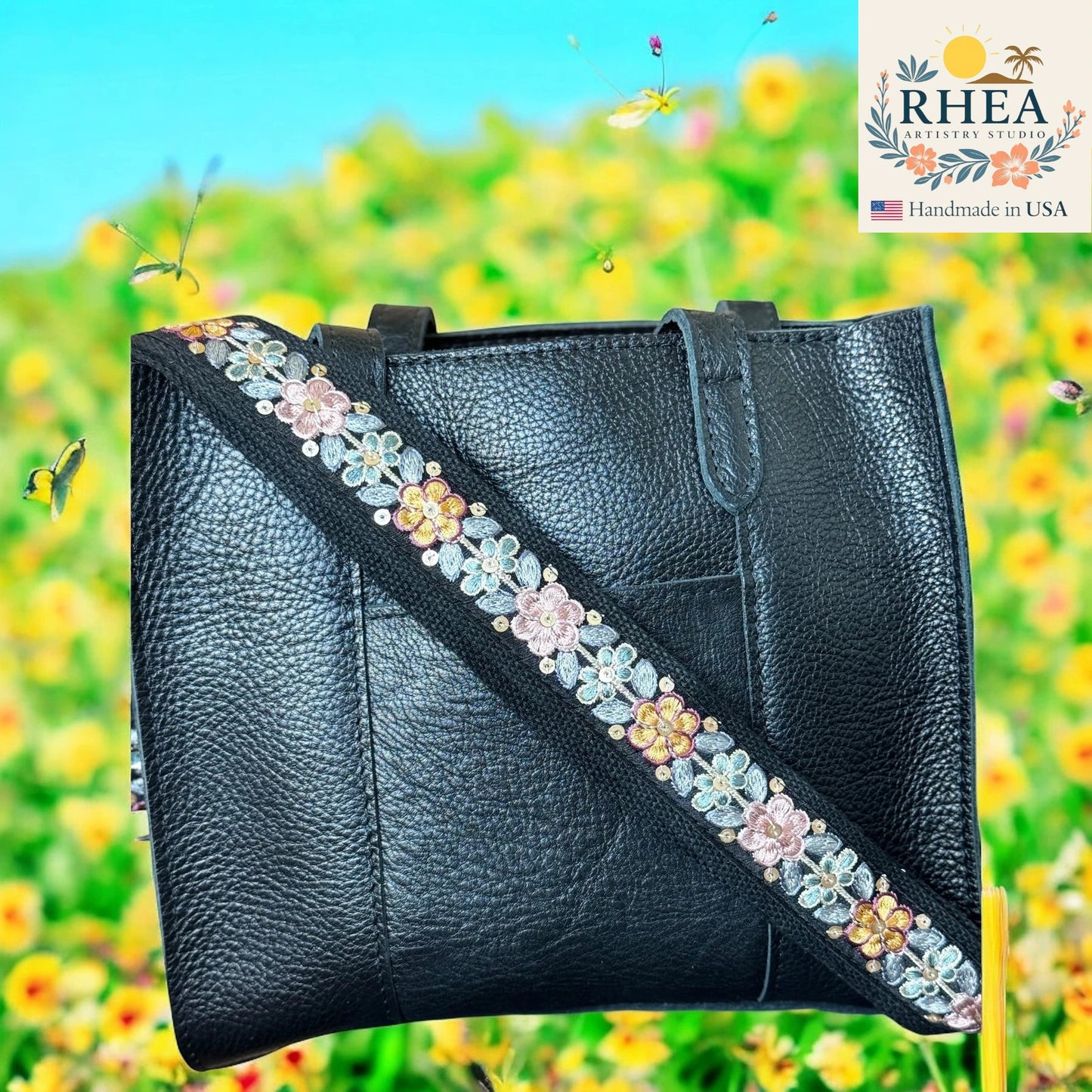 Black leather handbag with floral strap against a colorful floral background, featuring the RHEEA brand.