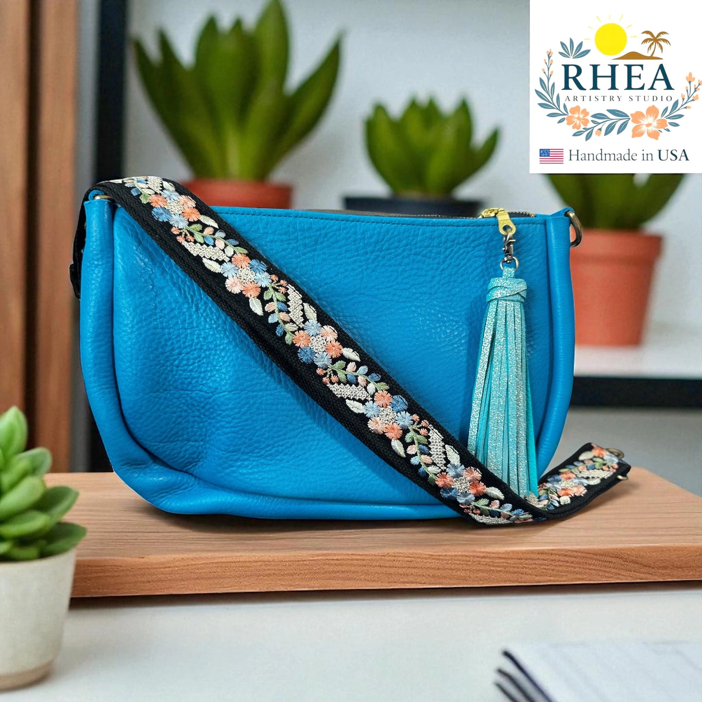 Blue handbag with decorative handmade artisan strap and tassel on a wooden surface, with 'RHEA' branding.