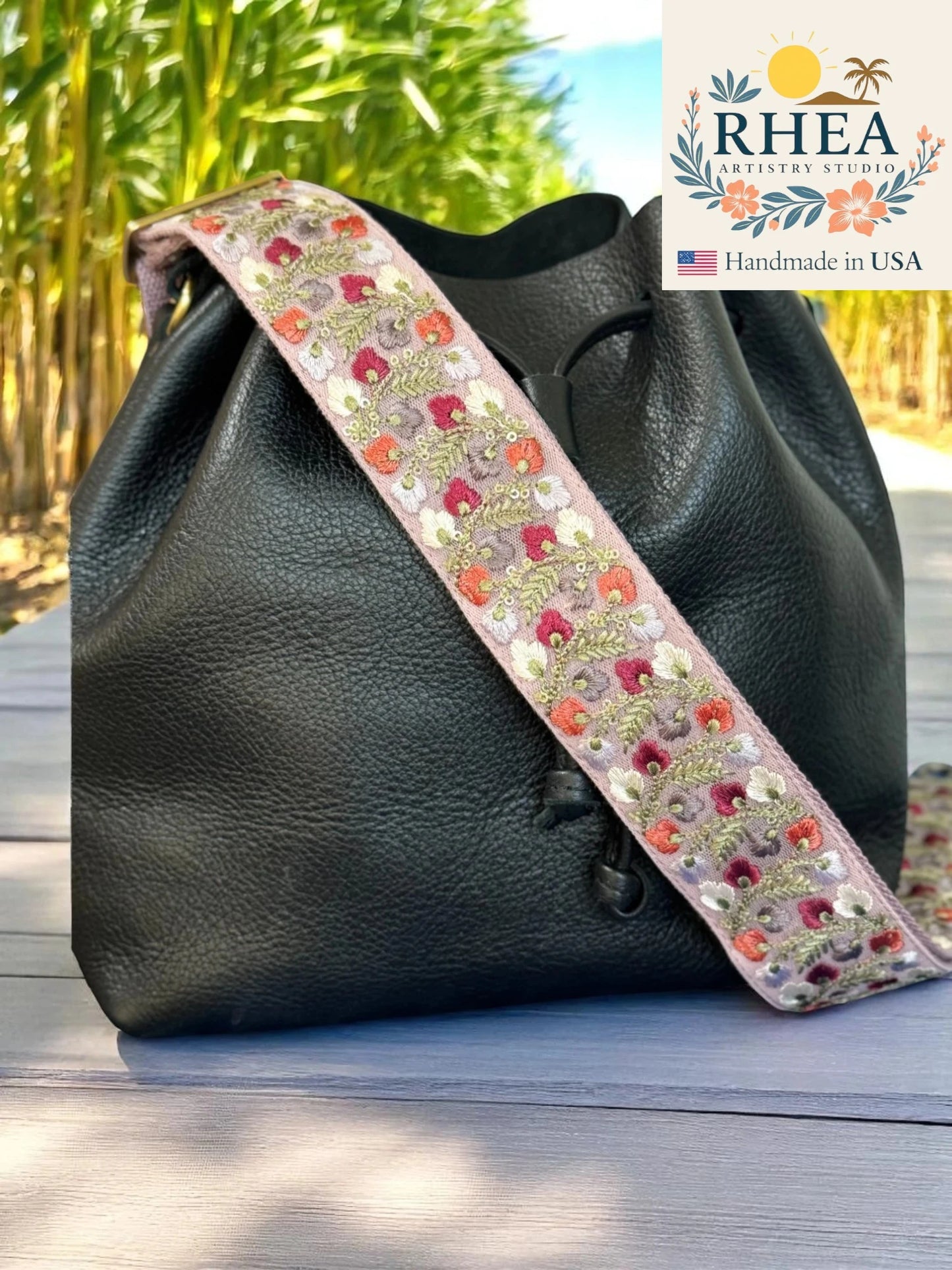 Black leather bag with a floral bag strap on a wooden surface, RHEA brand visible.