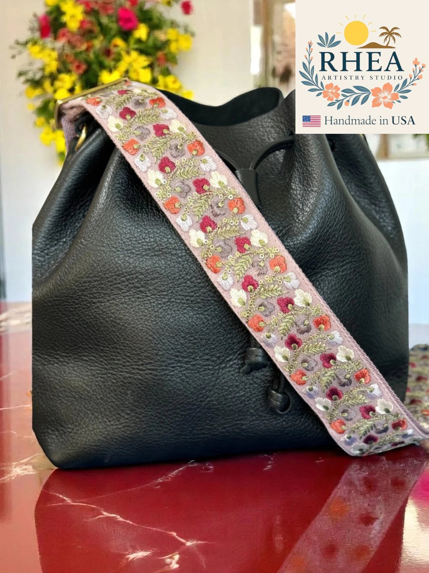 Black leather bag with a colorful floral purse strap on a reflective surface, with 'RHEA' branding.