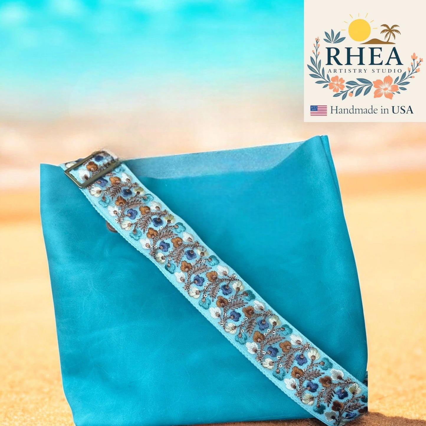 Turquoise fabric with floral embroidery bag strap on a beach background, featuring RHEA Artisan Studio logo.