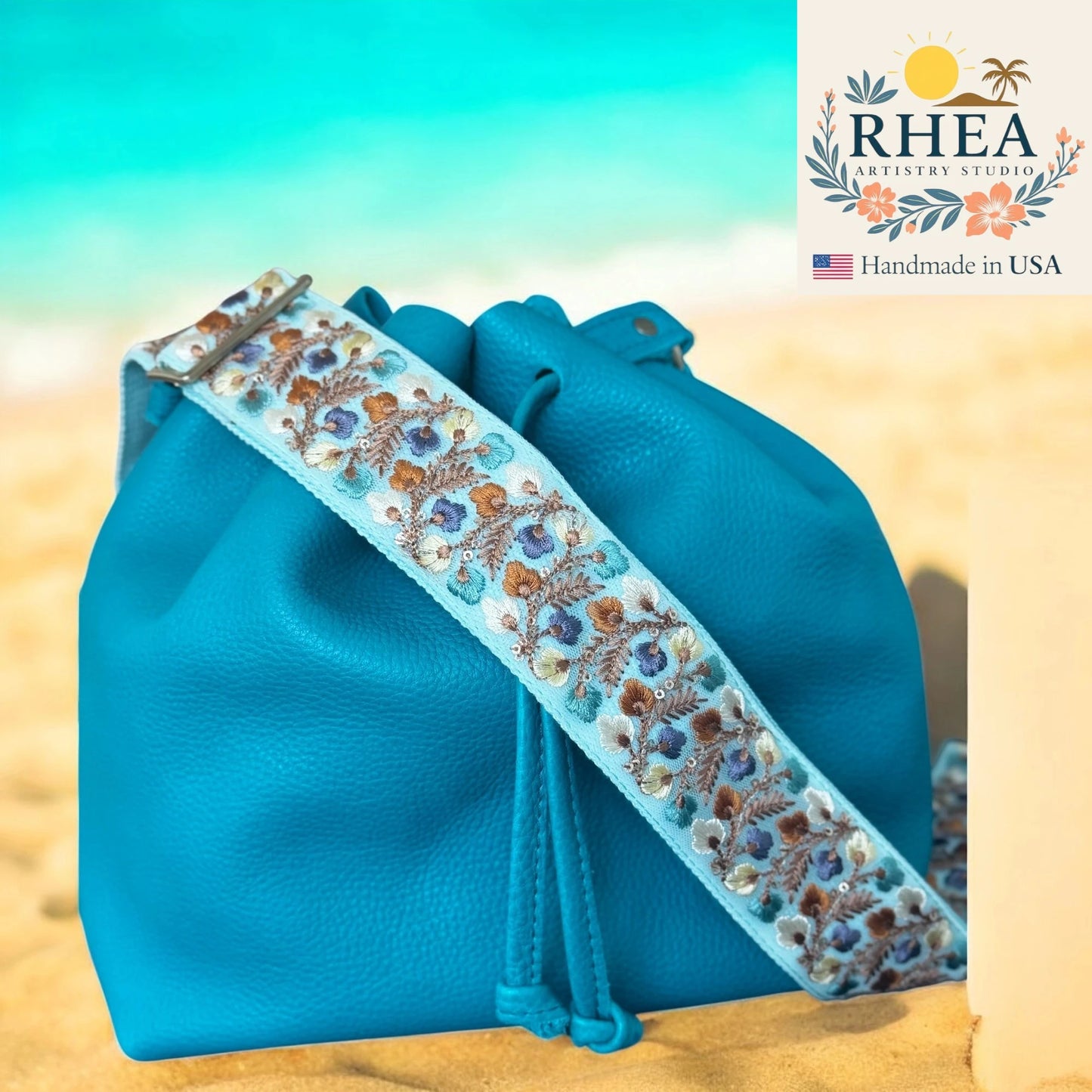 Blue drawstring bag with floral purse strap on a beach background, featuring the RHEA Artistry Studio logo.