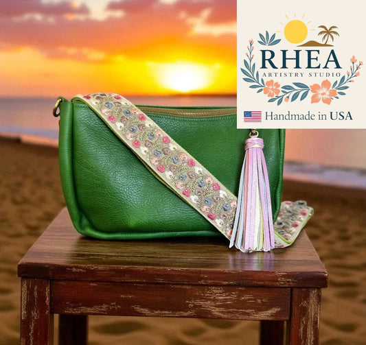 Green handbag with decorative purse strap and tassel on a wooden stool against a sunset beach backdrop, featuring RHEA Artistry Studio logo.
