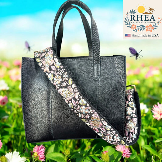 Black handbag with an embroidered purse strap against a floral background, featuring the RHEA brand.